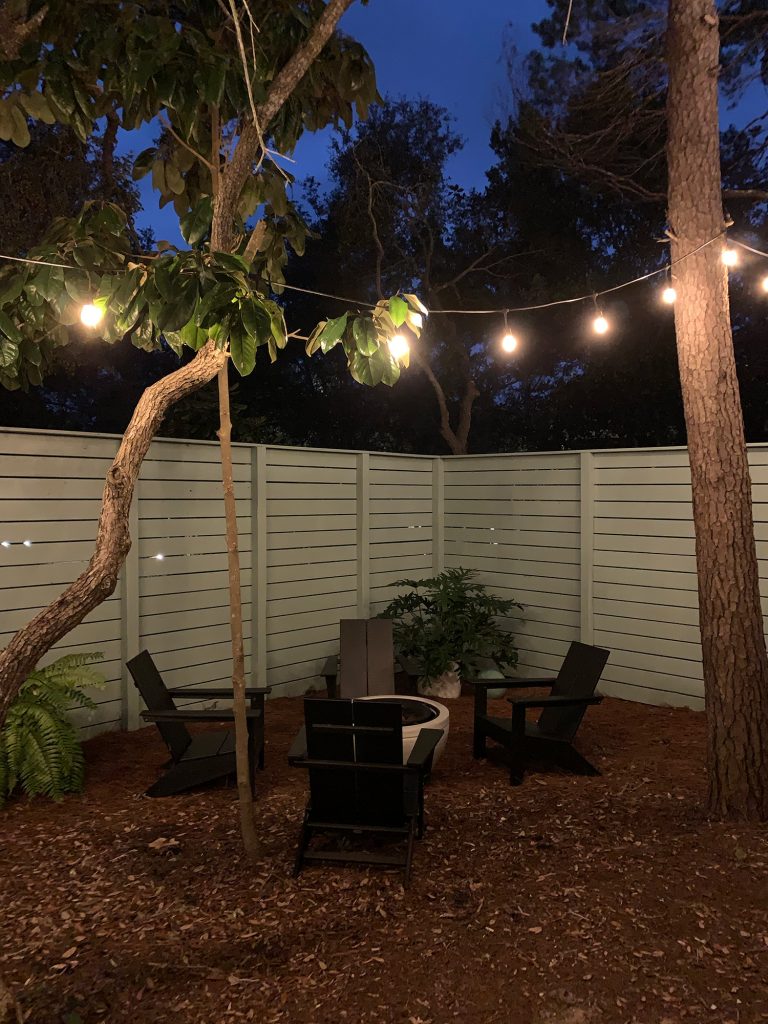 Nighttime view of fenced side yard with black Adirondack chairs around fire pit
