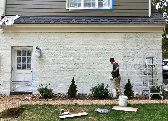 painter rolling a second coat of white paint on a brick house